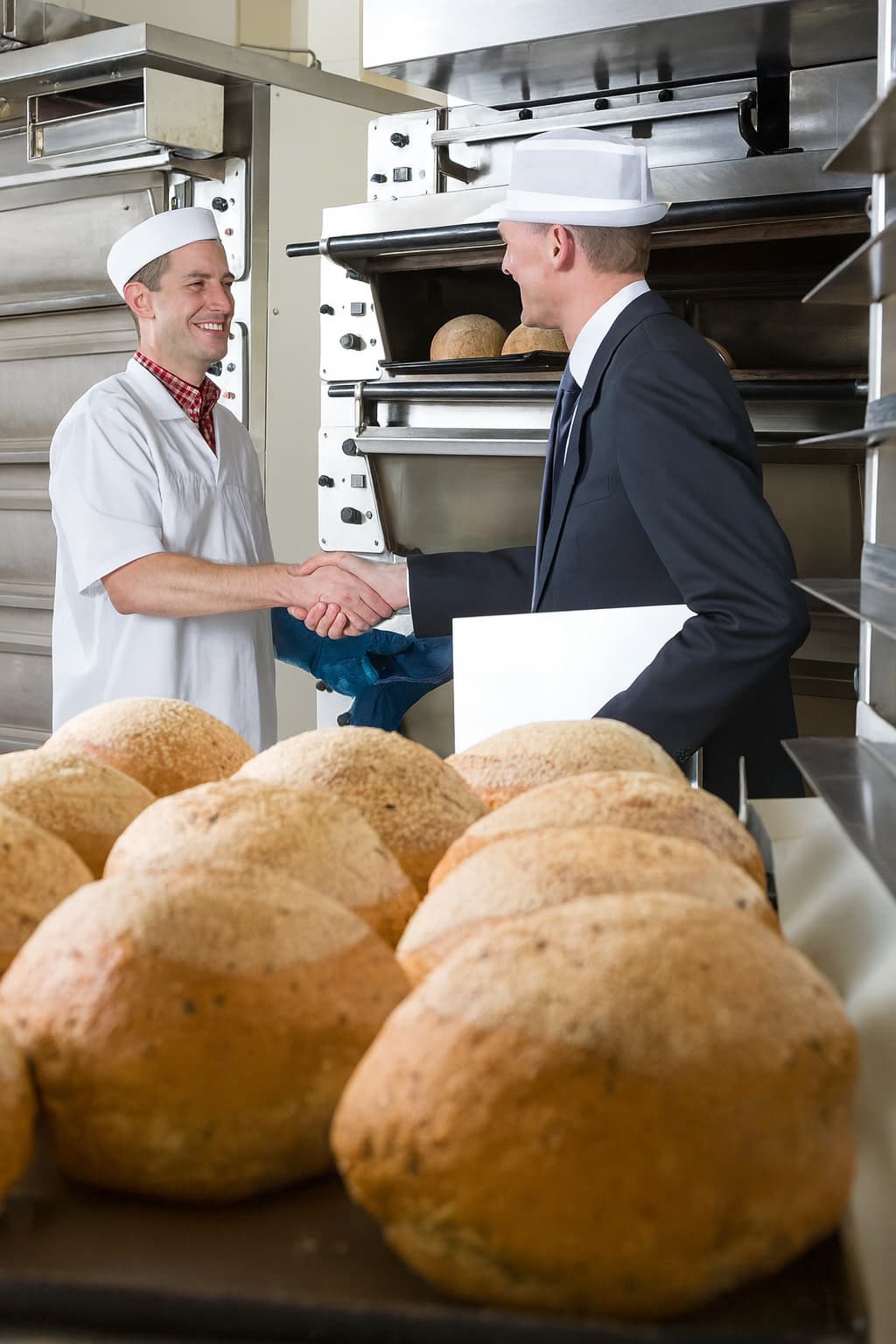 Baker and business professional shaking hands in a commercial bakery with trays of fresh bread rolls.