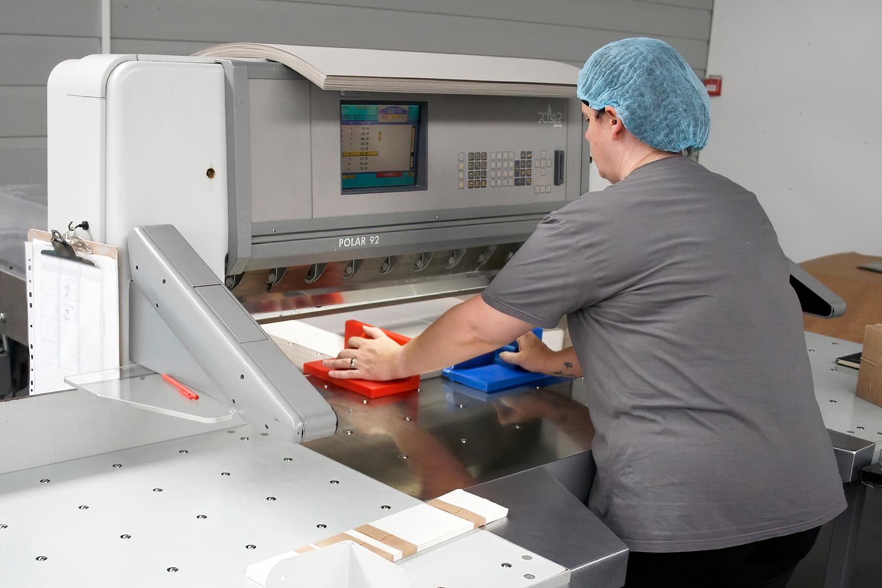 Production worker wearing a hairnet operates a Polar 92 industrial paper-cutting machine in a manufacturing facility.