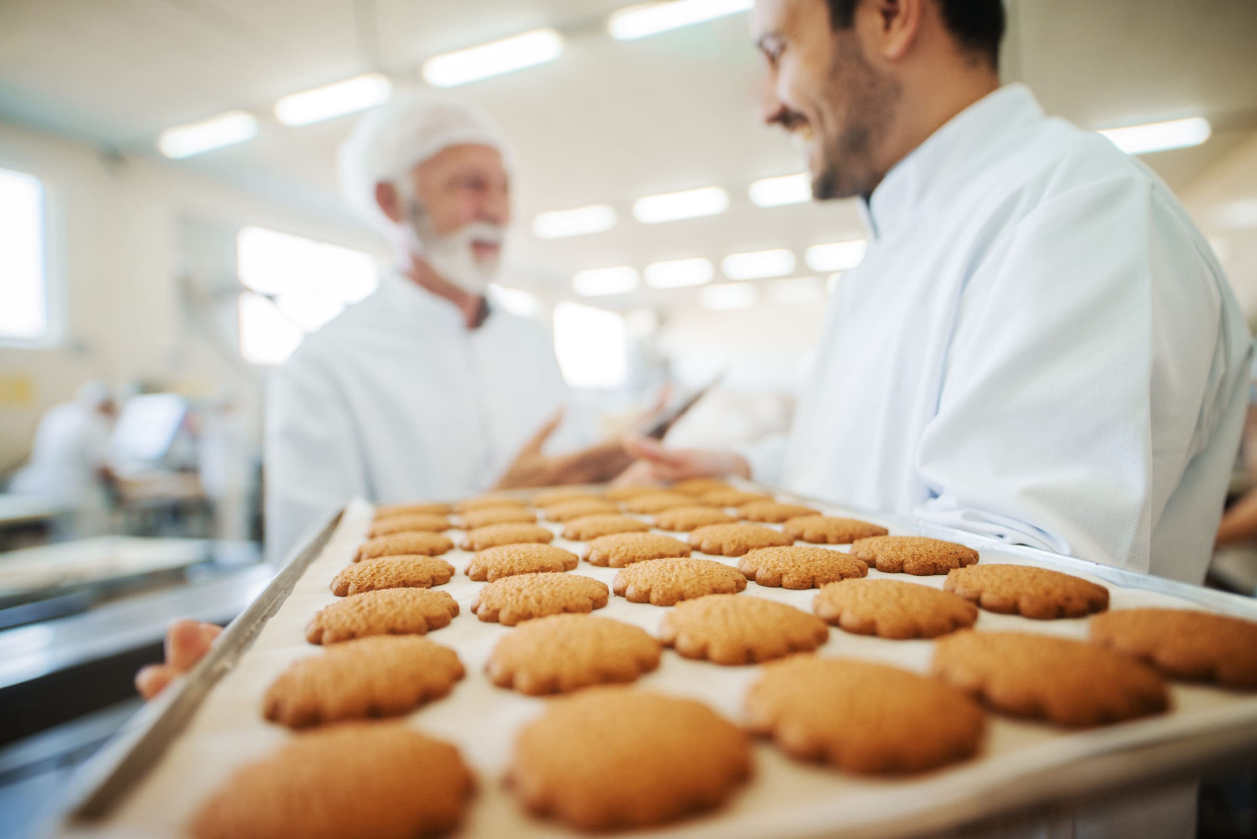 Bakery workers inspect freshly baked biscuits arranged on a paper-lined tray in a food production facility.