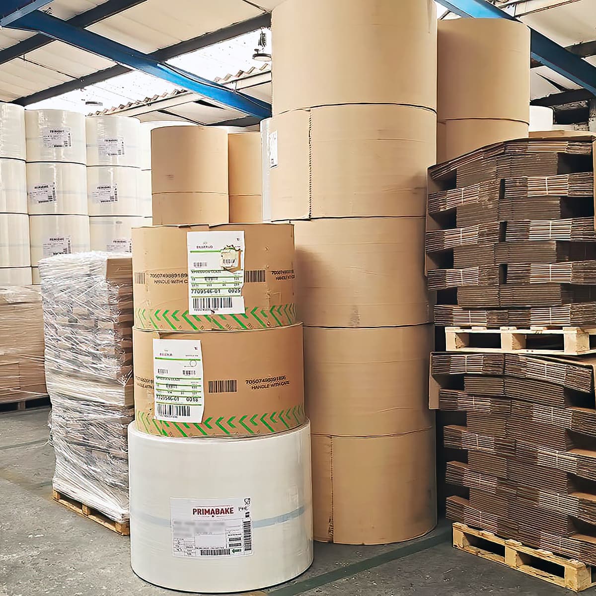 Large rolls of coated paper stored in a warehouse alongside packaged materials and stacks of flattened cardboard boxes on pallets.